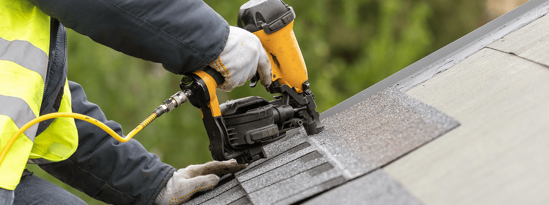 Worker using nail gun on roof shingles.